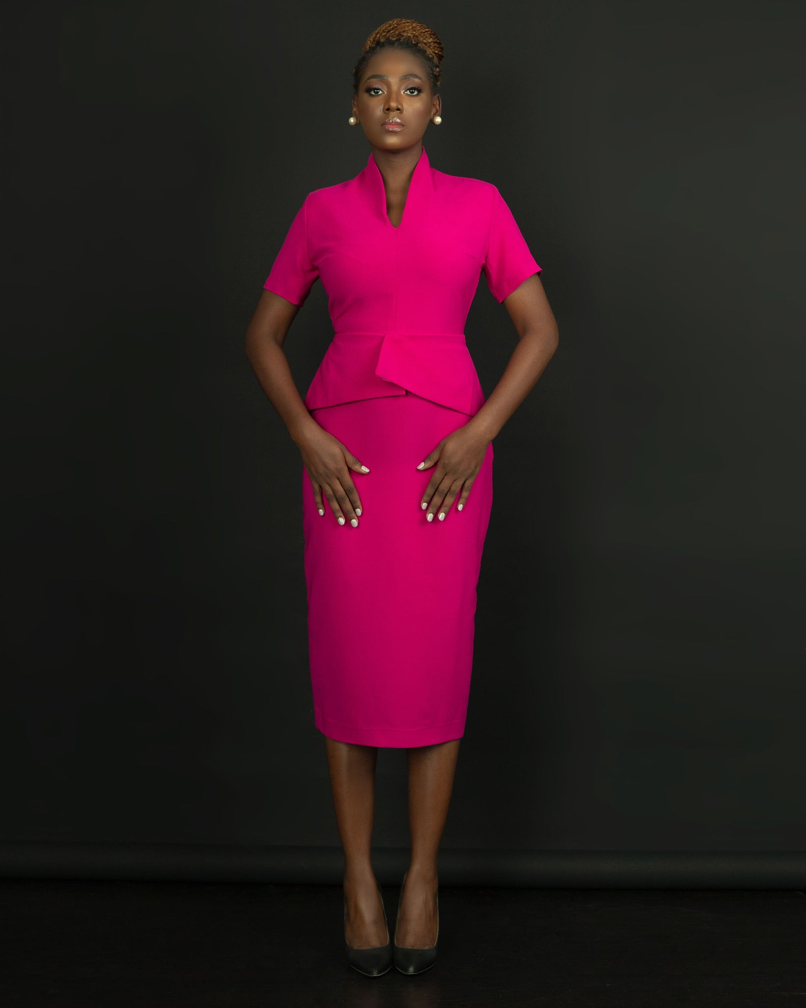 Woman in a fitted bright pink peplum dress, short sleeves, standing against a black background.