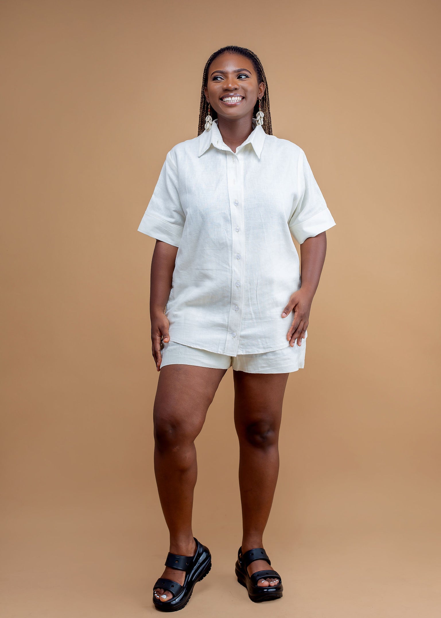 Smiling woman wearing a white linen shirt and shorts set, black sandals, studio background