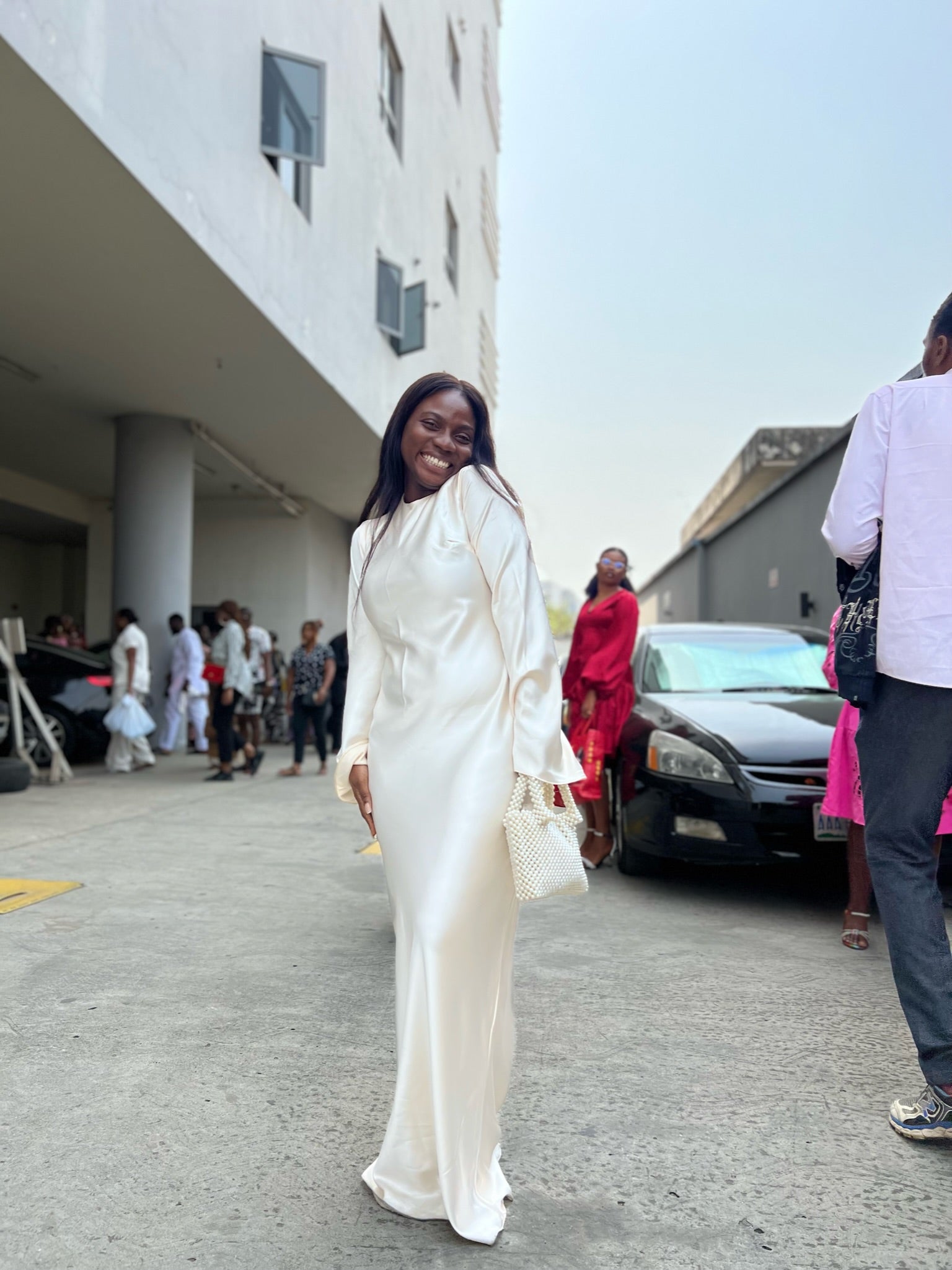 Woman in elegant white satin dress holding beaded bag, posing outdoors with a smile