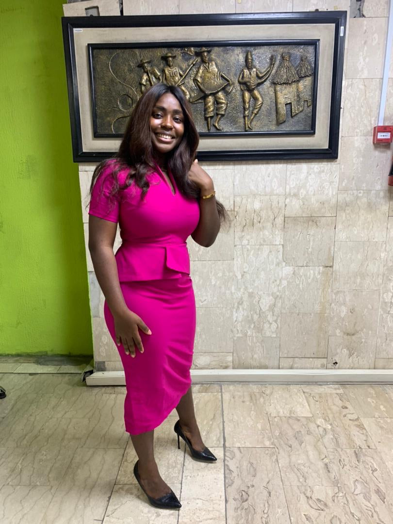 Smiling woman in a fitted pink dress and black heels standing indoors, with wall art behind her.