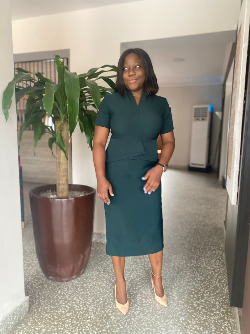 Woman in tailored green dress and nude heels standing indoors beside potted plant