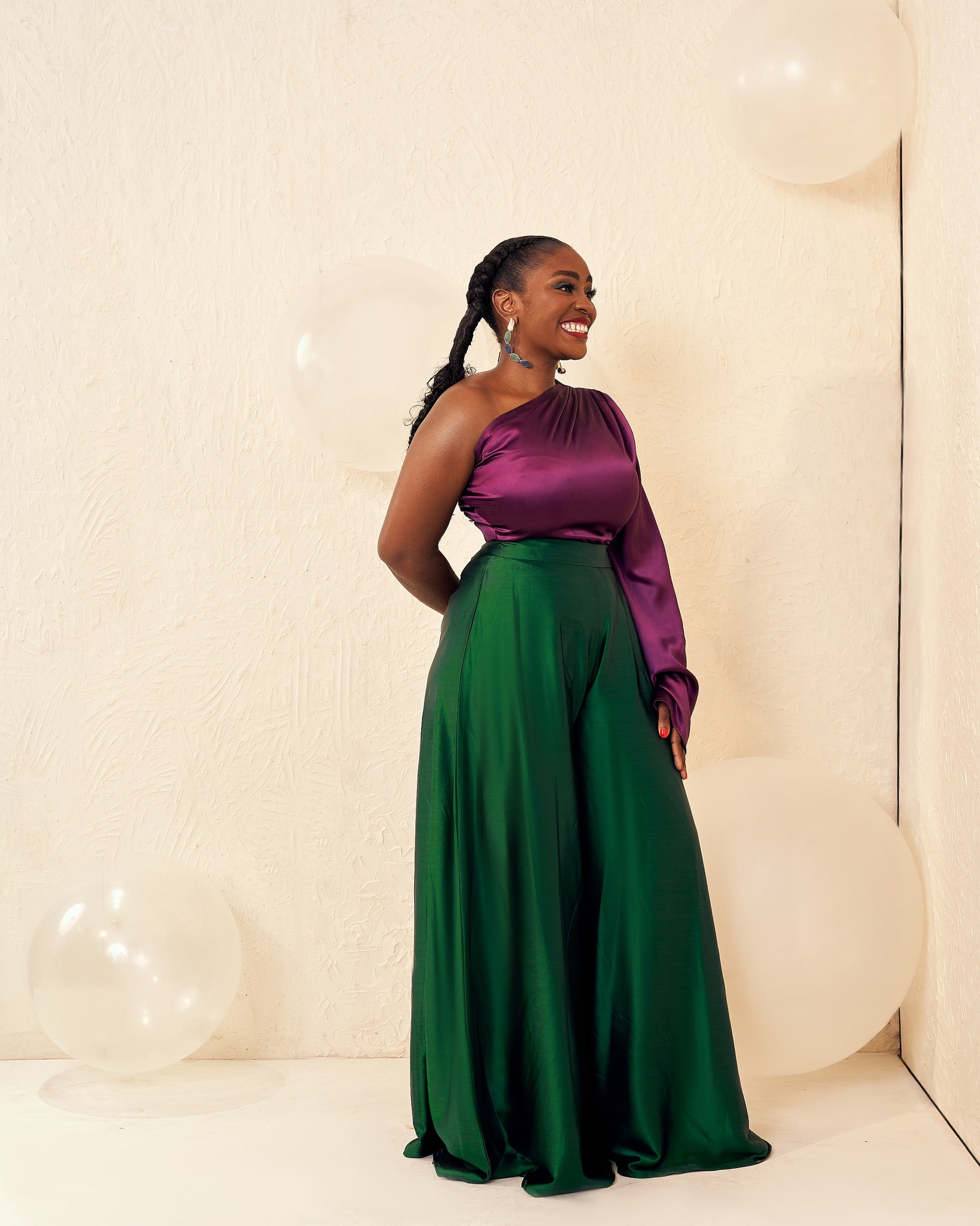 Smiling woman in elegant one-shoulder purple top and green wide-leg pants, standing by white balloons